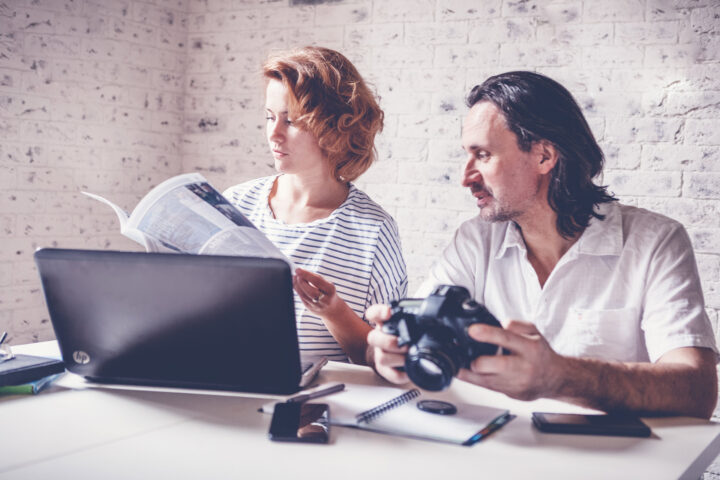 A middle-aged man and a young woman are sitting at a table with a laptop, camera and diary. Training and master classes in photography and processing, education concept, creative professions