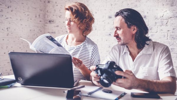 A middle-aged man and a young woman are sitting at a table with a laptop, camera and diary. Training and master classes in photography and processing, education concept, creative professions